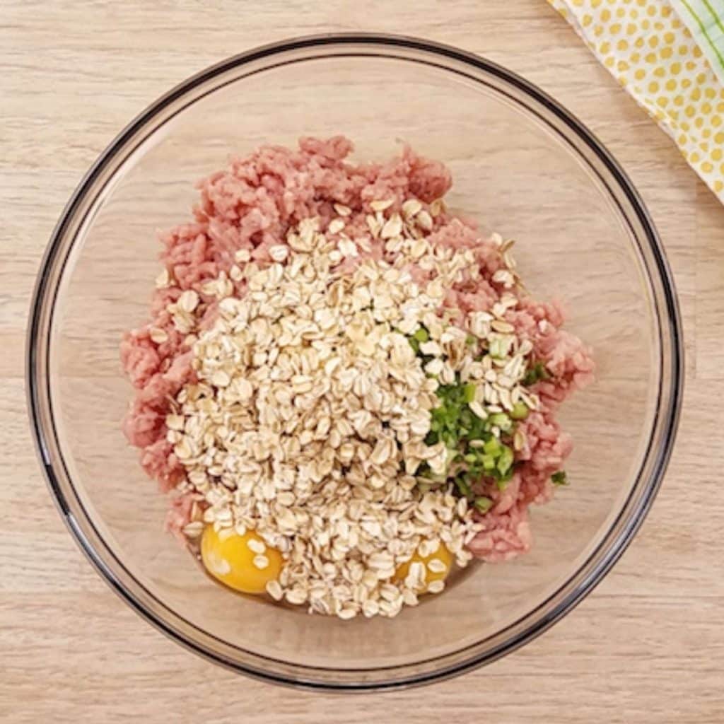 A glass bowl filled with ground turkey, eggs, seasonings and oats, ready to be mixed together to form meatballs.