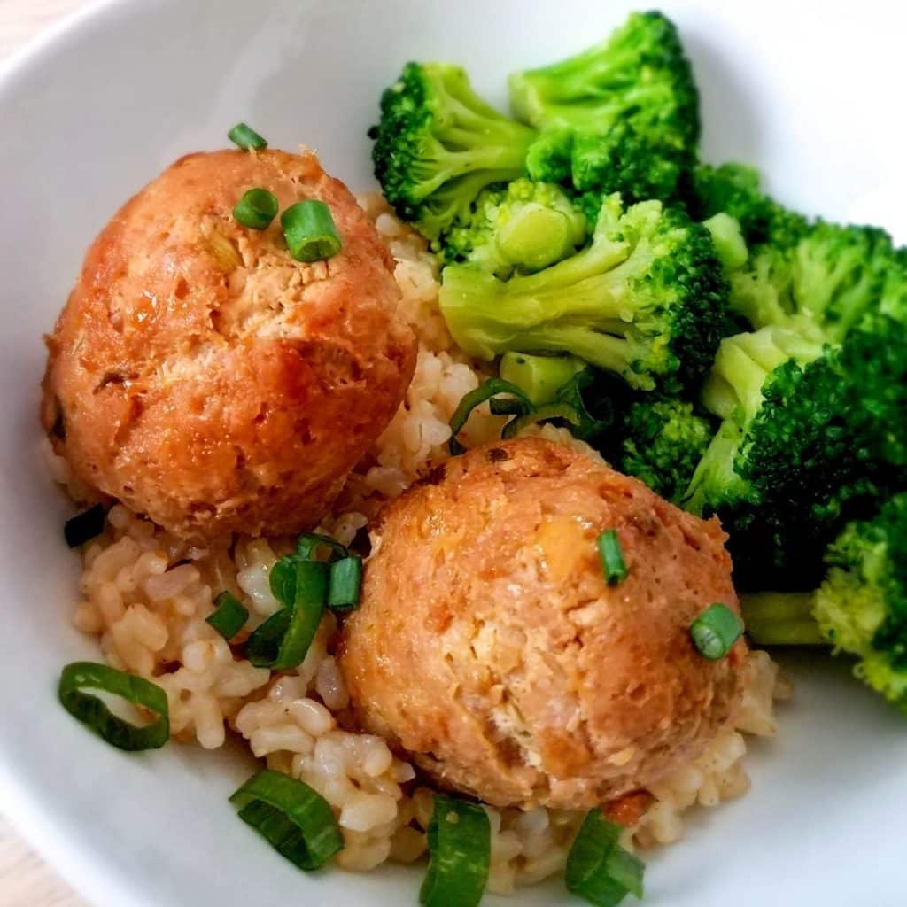 A white bowl with cooked brown rice, two teriyaki meatballs, broccoli and topped with green onions.