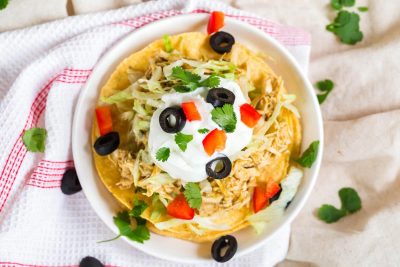 one tostada from above showing shredded green chili chicken, lettuce, sour cream, tomatoes, cilantro, and black olives
