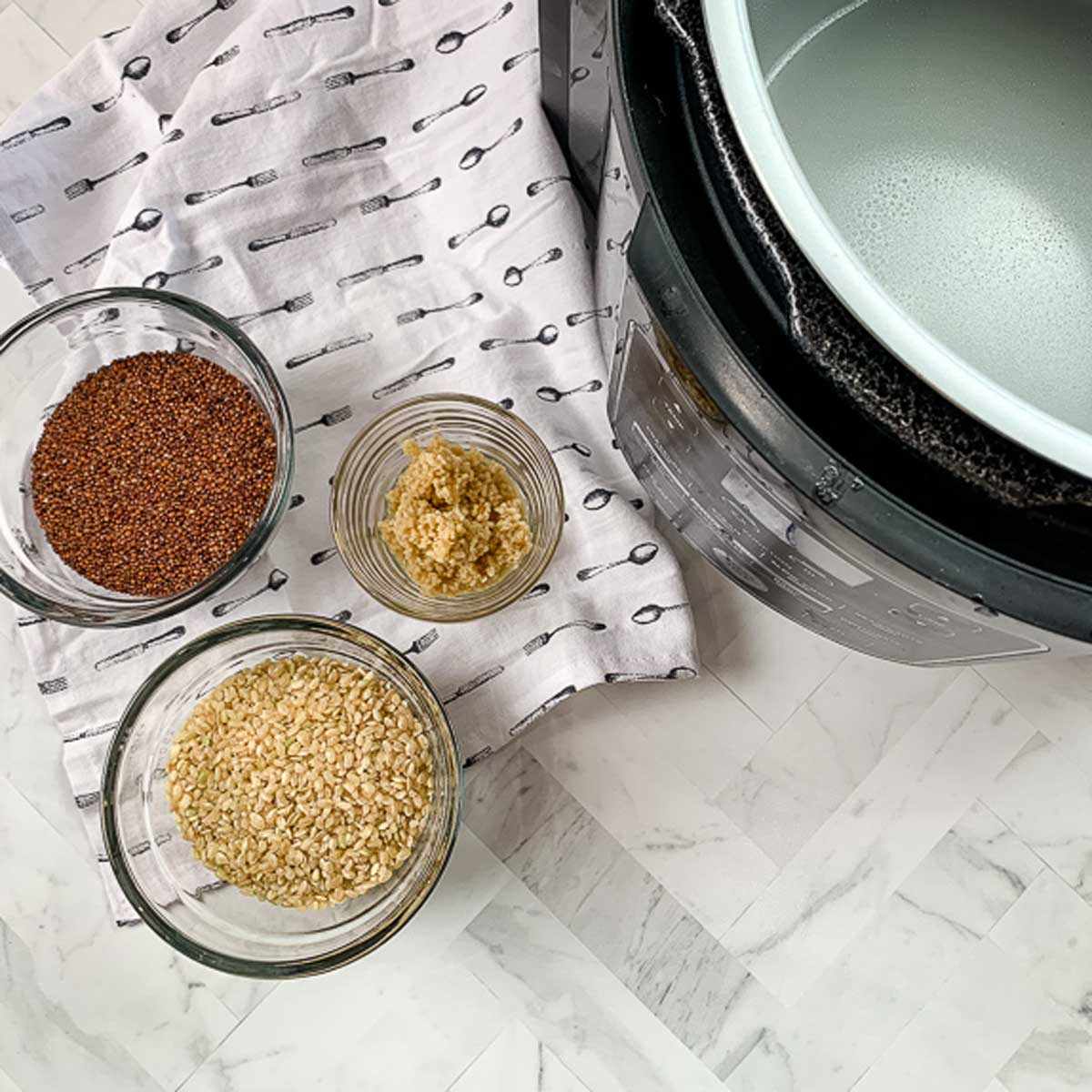 Three glass bowls sitting on top of a white and black napkin with each containing ingredients to make brown rice and quinoa. 