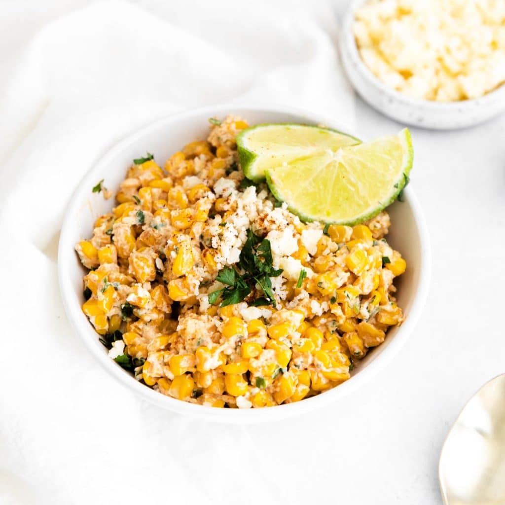 A white counter with a white bowl full of easy and healthy elote dip and topped with lime wedges and parsley. Off the the side is a spoon, a bowl of tortilla chips, another wedge of lime, and a bowl of spices.