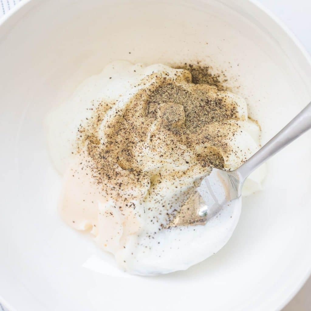A white bowl with mayonnaise, greek yogurt, vanilla extract, and pepper in it being mixed by a spoon.