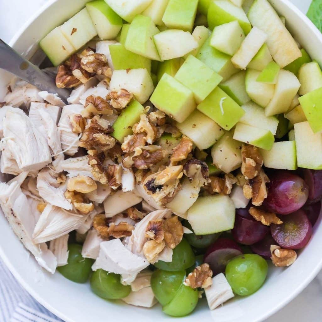 A white bowl with cut up green apples, diced chicken breast, walnut pieces, and cut green and red grapes.