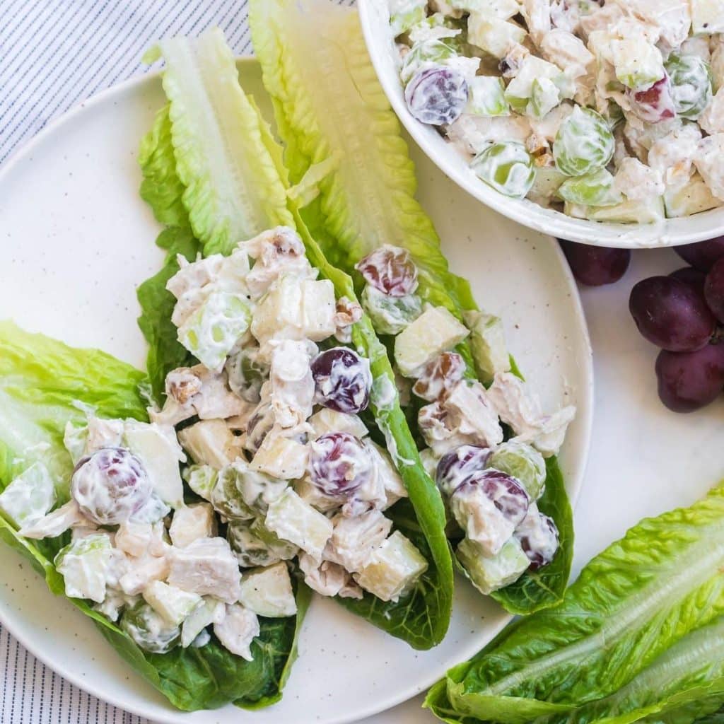 Three pieces of romaine lettuce on a white plate and topped with diced chicken that has been mixed together with a yogurt, grape and apple mixture.