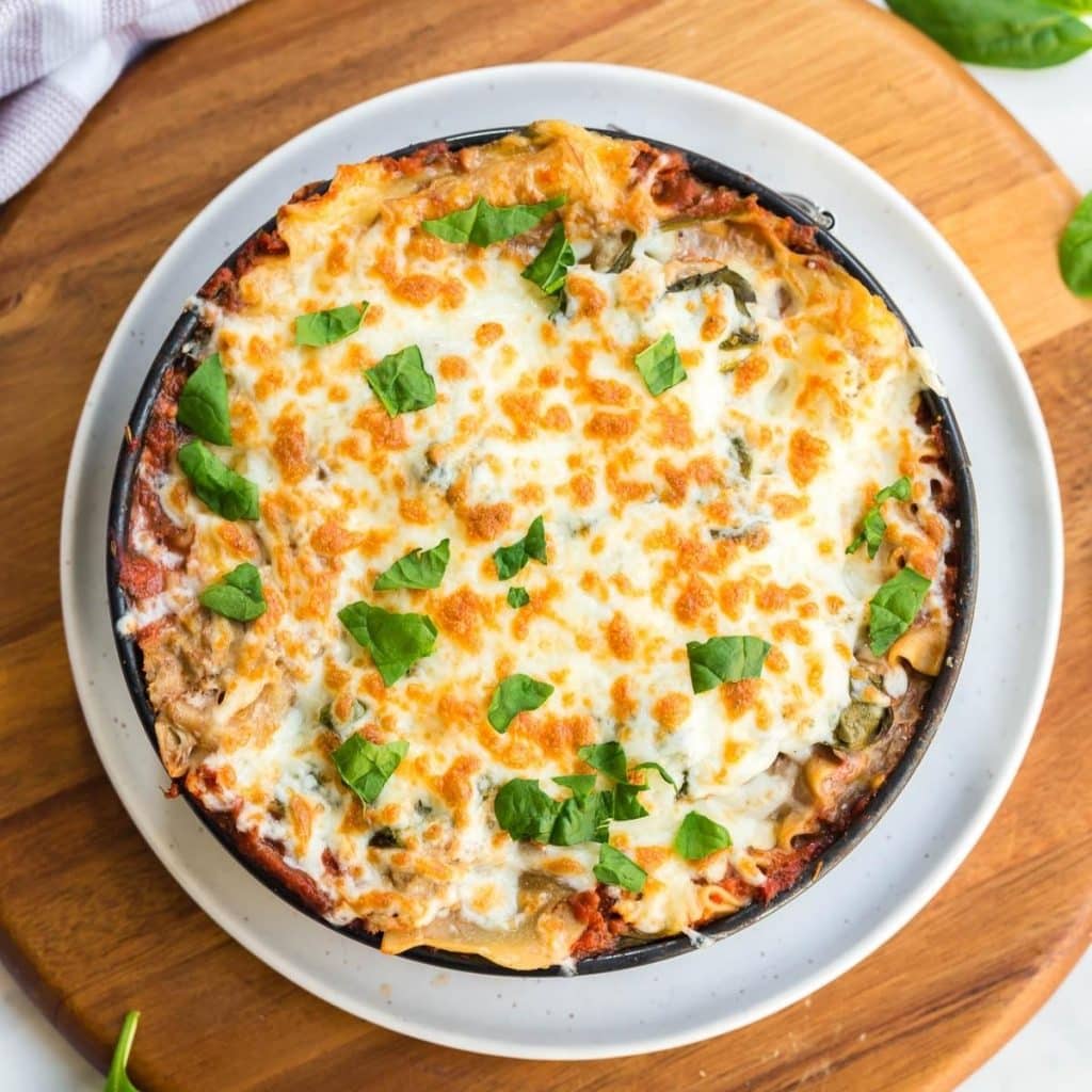 Cooked lasagna in a circular baking dish showing melted cheese topped with chopped basil.