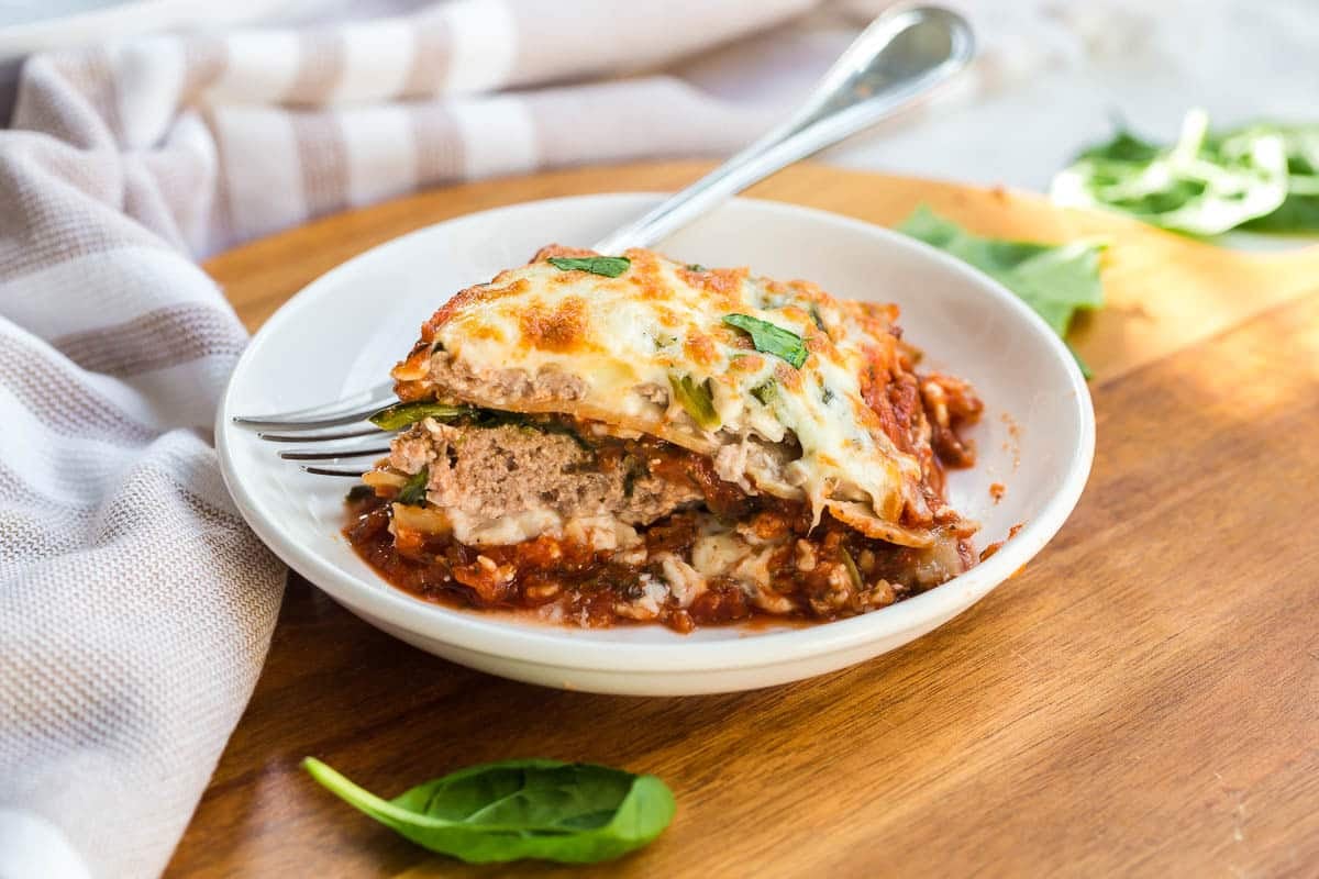 A wooden board with a white bowl on top that has a slice of healthy lasagna in it.