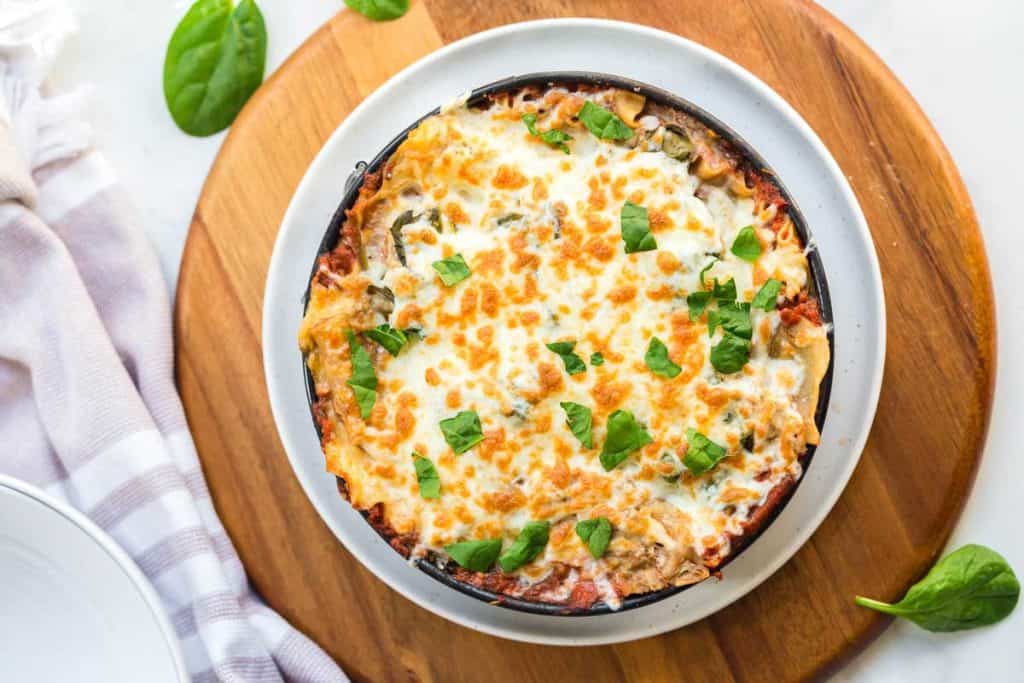 Image of full lasagna recipe on a white plate on top of a wooden platter. The lasagna is garnished with fresh basil leaves. Cheese is melted and slightly browned. 