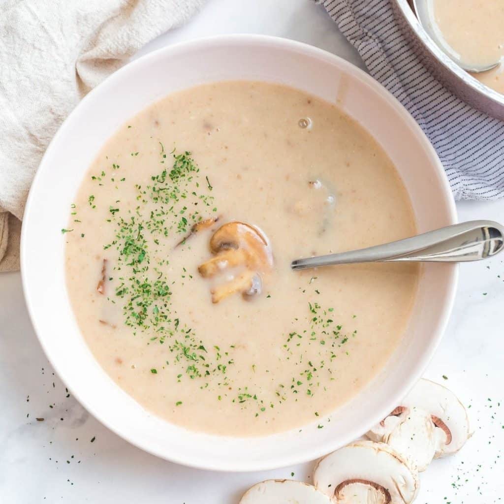A white bowl with homemade cream of mushroom soup that shows a sliced mushroom and a scattering of green parsley flakes on top.