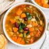 cooked minestrone soup in white bowl with crusty bread and napkin on counter