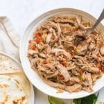 White countertop with a white bowl of shredded pork carnitas and a white plate of flour tortillas off to the left.