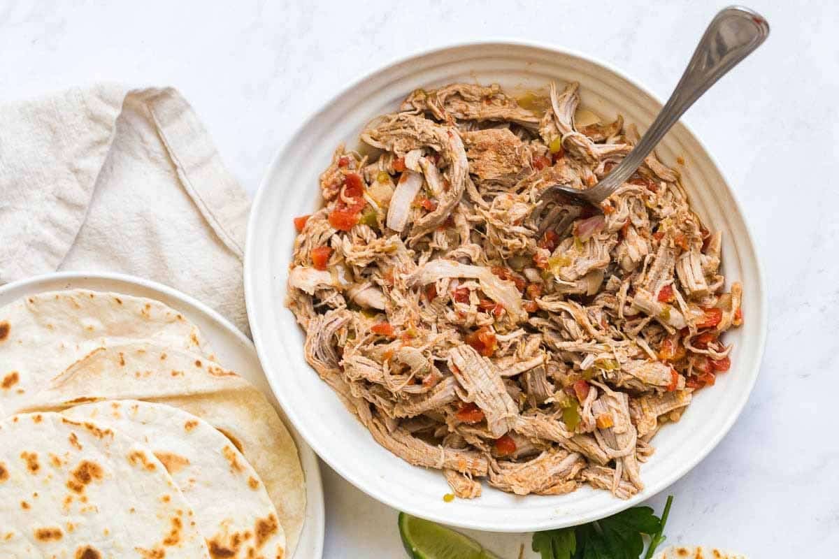 White countertop with a white bowl of shredded pork carnitas and a white plate of flour tortillas off to the left. 