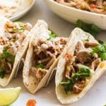 Marble counter with three carnitas tacos with toppings, a white plate of flour tortillas, and a white bowl of carnitas meat.