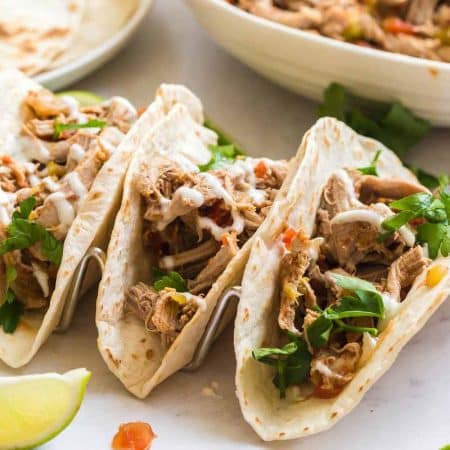 Marble counter with three carnitas tacos with toppings, a white plate of flour tortillas, and a white bowl of carnitas meat.