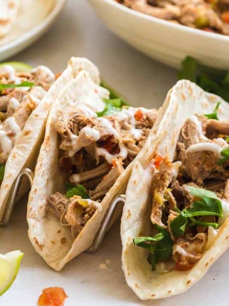 Marble counter with three carnitas tacos with toppings, a white plate of flour tortillas, and a white bowl of carnitas meat.