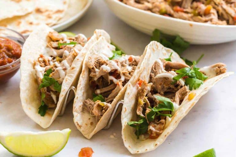 Marble counter with three carnitas tacos with toppings, a white plate of flour tortillas, and a white bowl of carnitas meat.