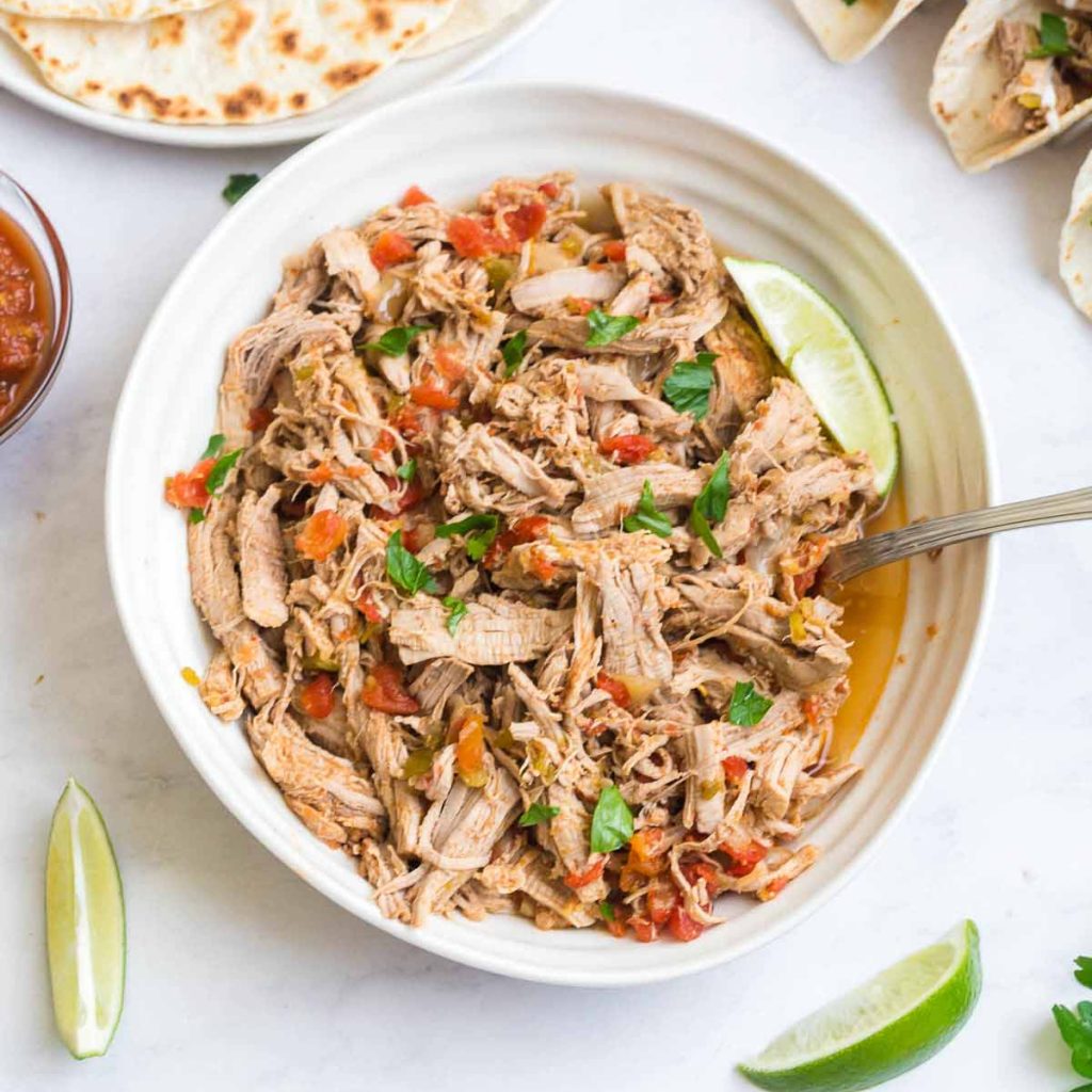 Large white bowl of carnitas with ime garnish on a white countertop. There are limes and parsley around the background. 