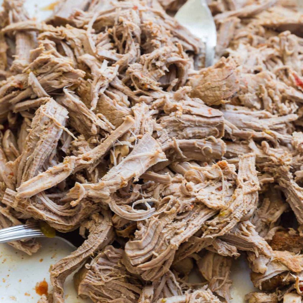 White plate with pork being shredded by two forks.