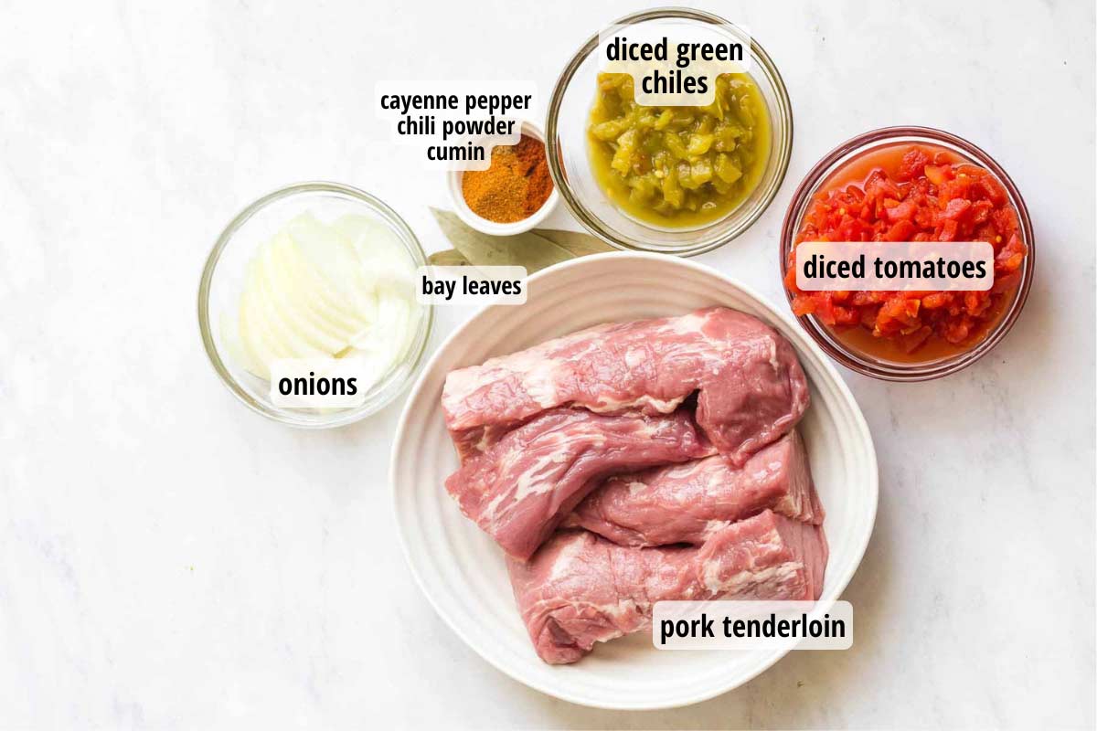 Marble counter with ingredients in clear and white bowls: pork tenderloin, onions, spices, green chiles, and diced tomatoes.