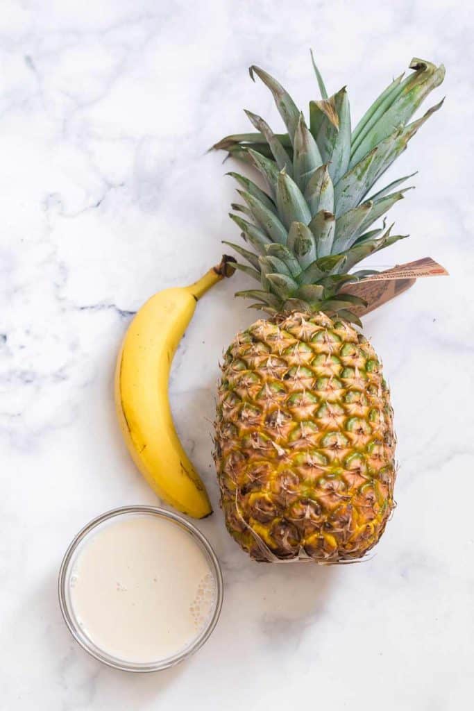 milk in a clear glass bowl, whole banana, and a whole pineapple on a white countertop