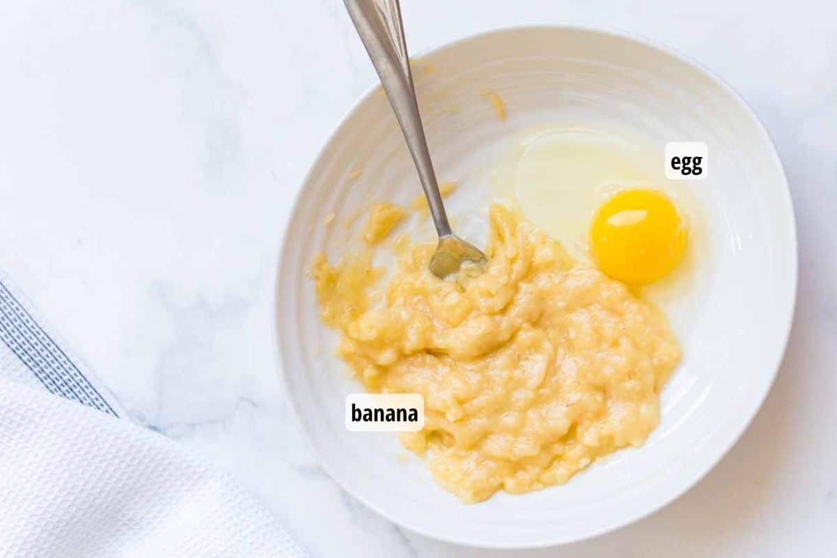 A white bowl on top of a white marbled counter with a mashed banana and egg about to be mixed together with a fork.