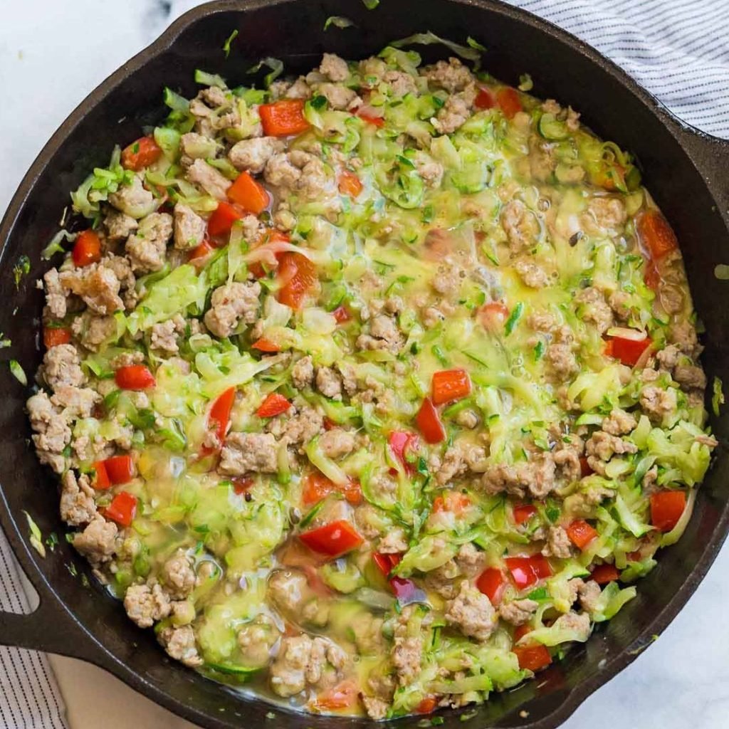 cast iron skillet with cooked sausage and zucchini and scrambled uncooked egg on top