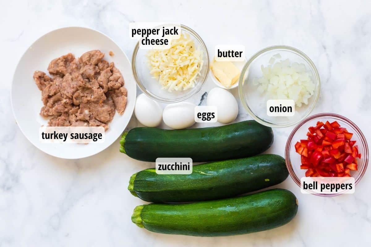 A white marbled counter top with bowls of labeled ingredients of sausage, cheese, onion and bell pepper. Within the image there are also 3 zucchinis and 3 eggs.
