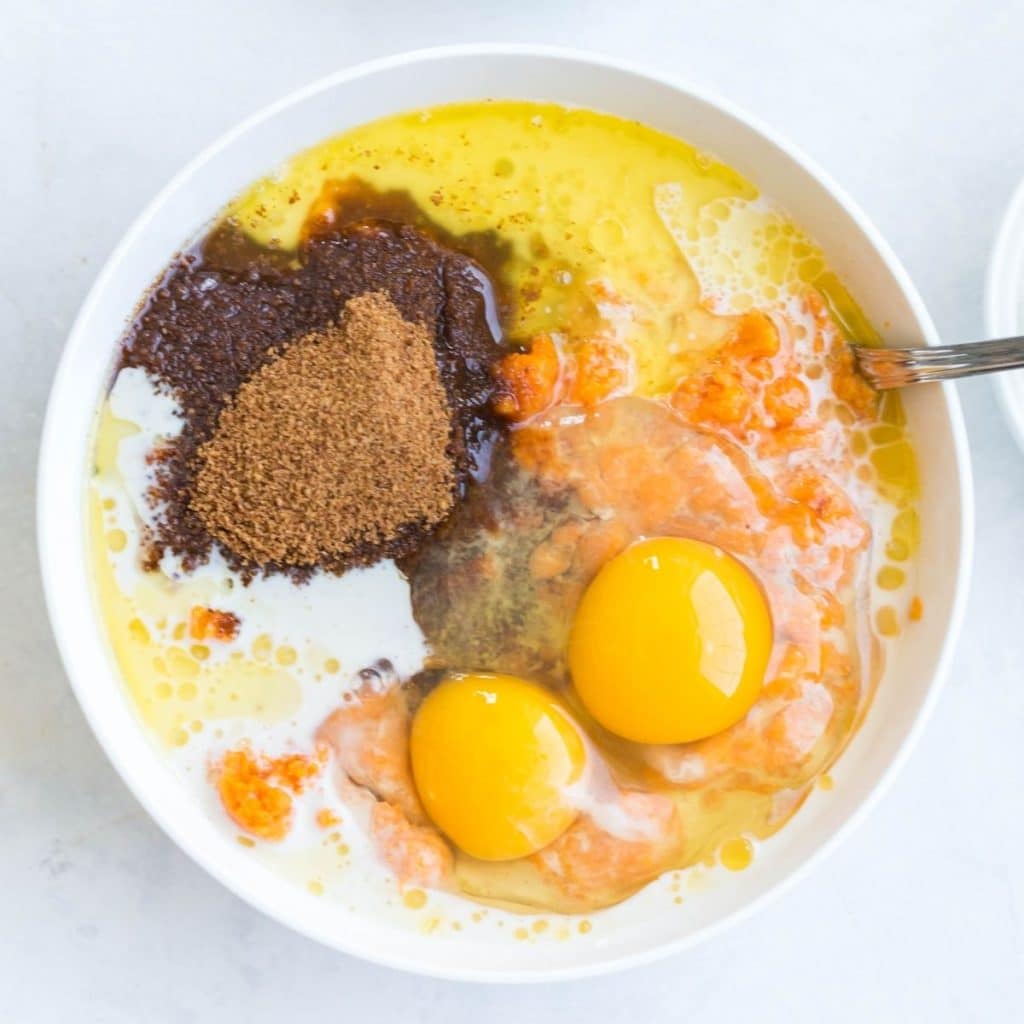A white bowl with ingredients like eggs, mashed sweet potatoes, coconut sugar, coconut milk and butter all about to be mixed together.