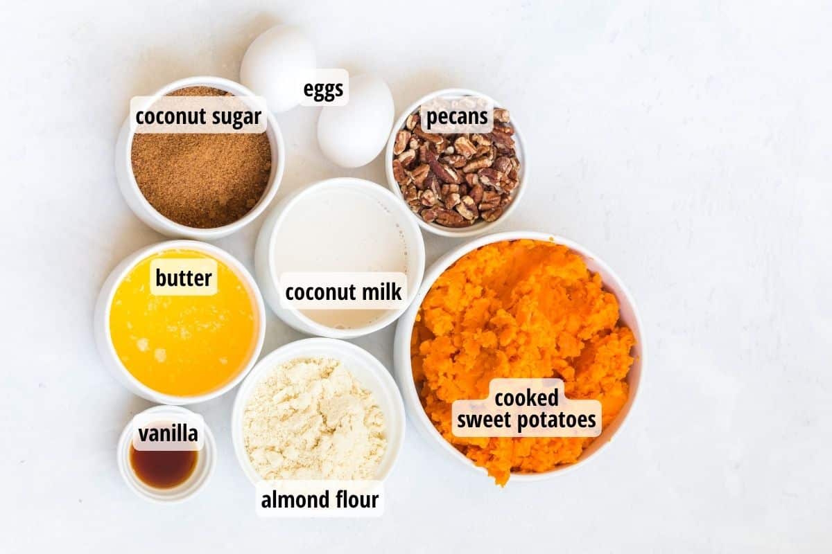 A white counter top with two eggs and seven white bowls filled with labeled ingredients needed to make healthy sweet potato casserole.