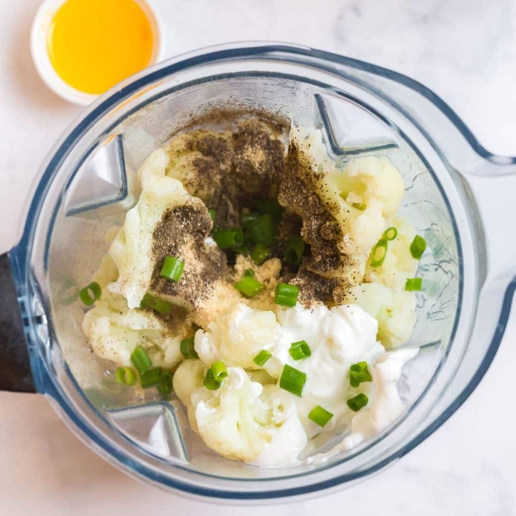 A blender with steamed cauliflower, greek yogurt, spices, ghee and green onions, all about to be blended together to make mashed cauliflower.