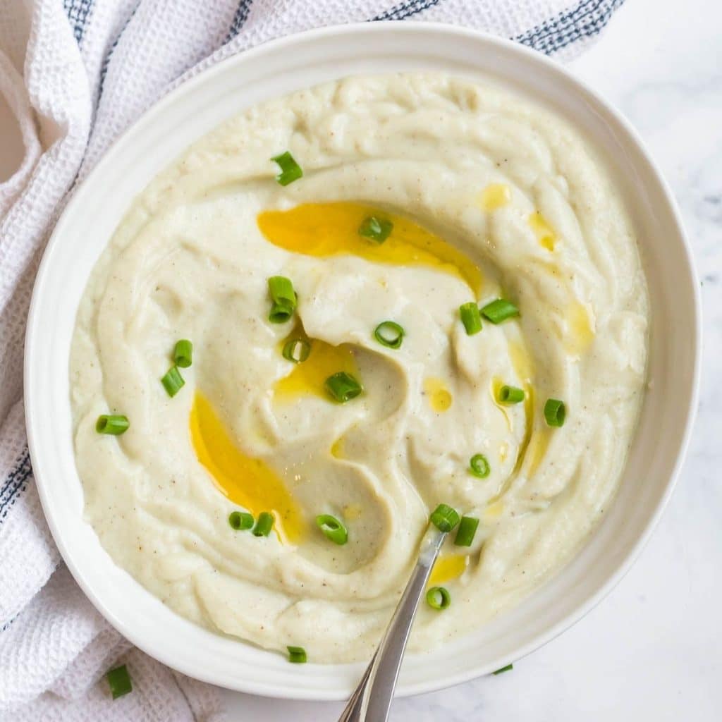 Mashed cauliflower topped with chives and a drizzle of ghee inside of a white bowl which is sitting on a white linen napkin.