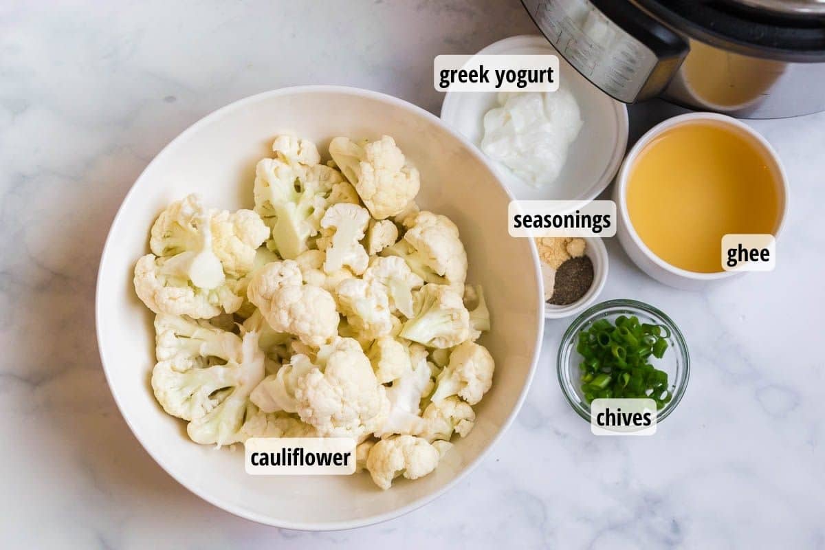 A white marbled counter top with bowls of labeled ingredients to include cauliflower florets, chives, seasonings, greek yogurt, and ghee.
