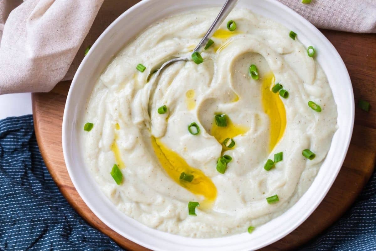 Mashed cauliflower topped with chives and a drizzle of ghee inside of a white bowl which is sitting on a wooden circular board. 