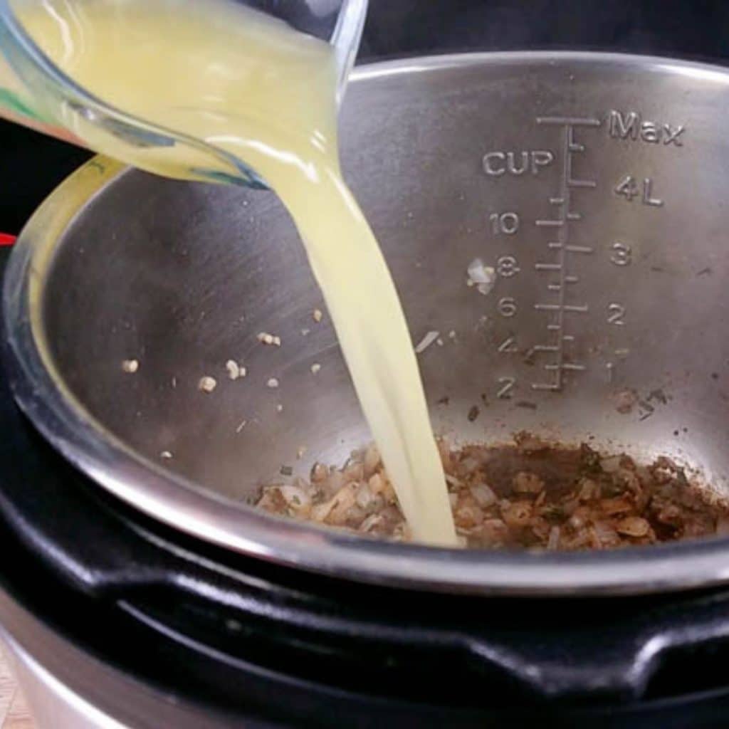 A glass measuring cup pouring broth into an instant pot with onion, garlic and herbs inside.