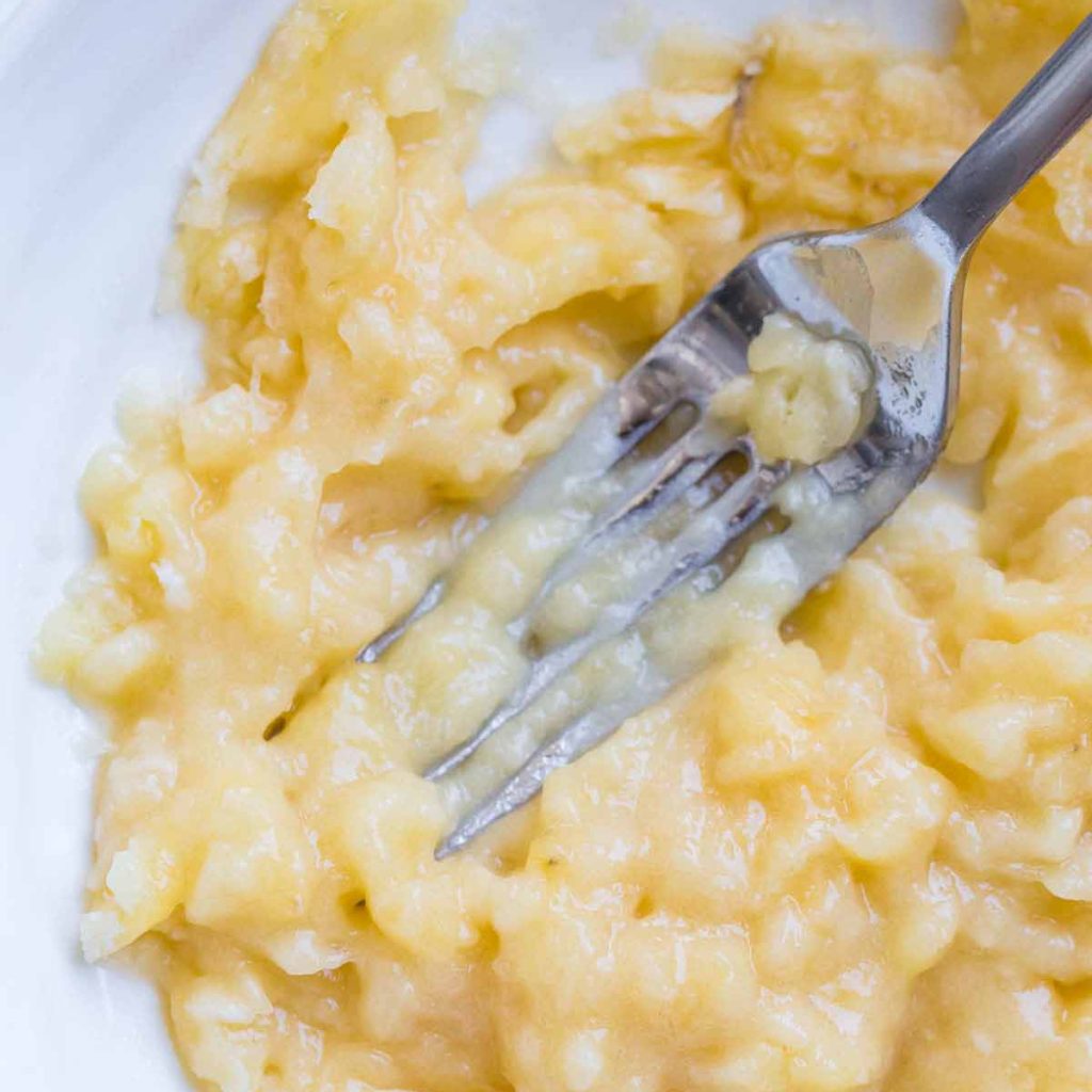 Fork tines mashing a banana inside of a white bowl.