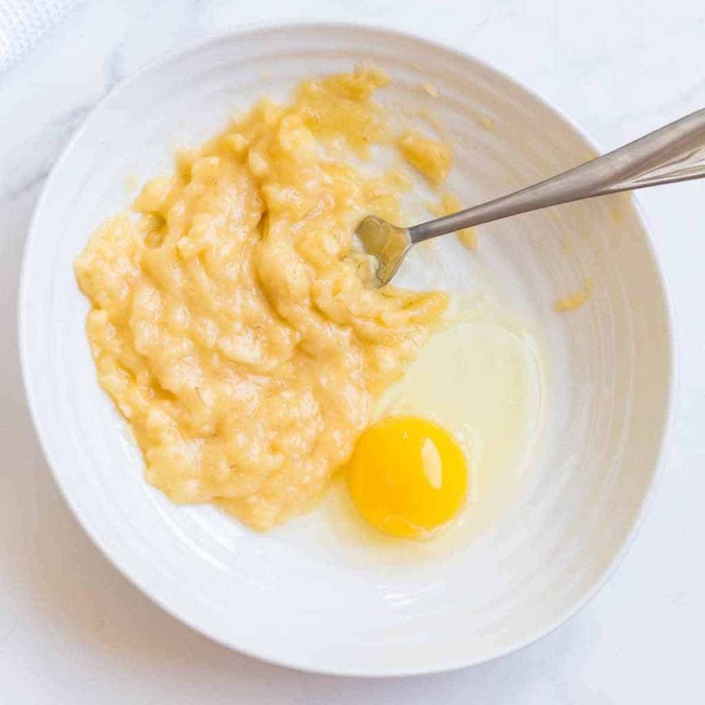 White bowl with a mashed banana and a whole egg inside. Fork is off to the side of the bowl.