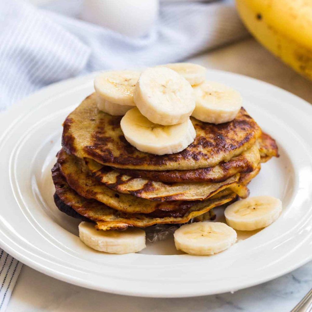 Stack of banana pancakes on a white plate with sliced bananas on top and around the bottom of the stack.