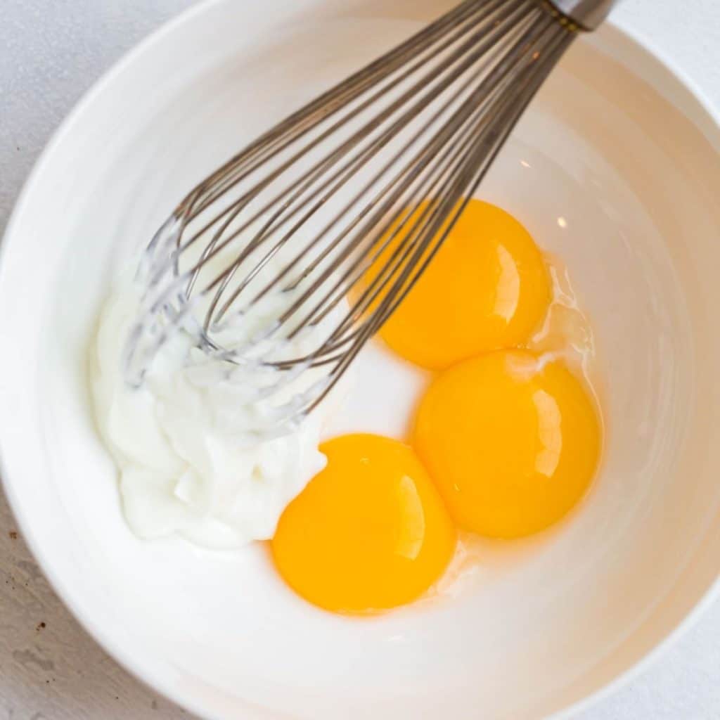 A white bowl with plain greek yogurt and three egg yolks being mixed together with a whisk.