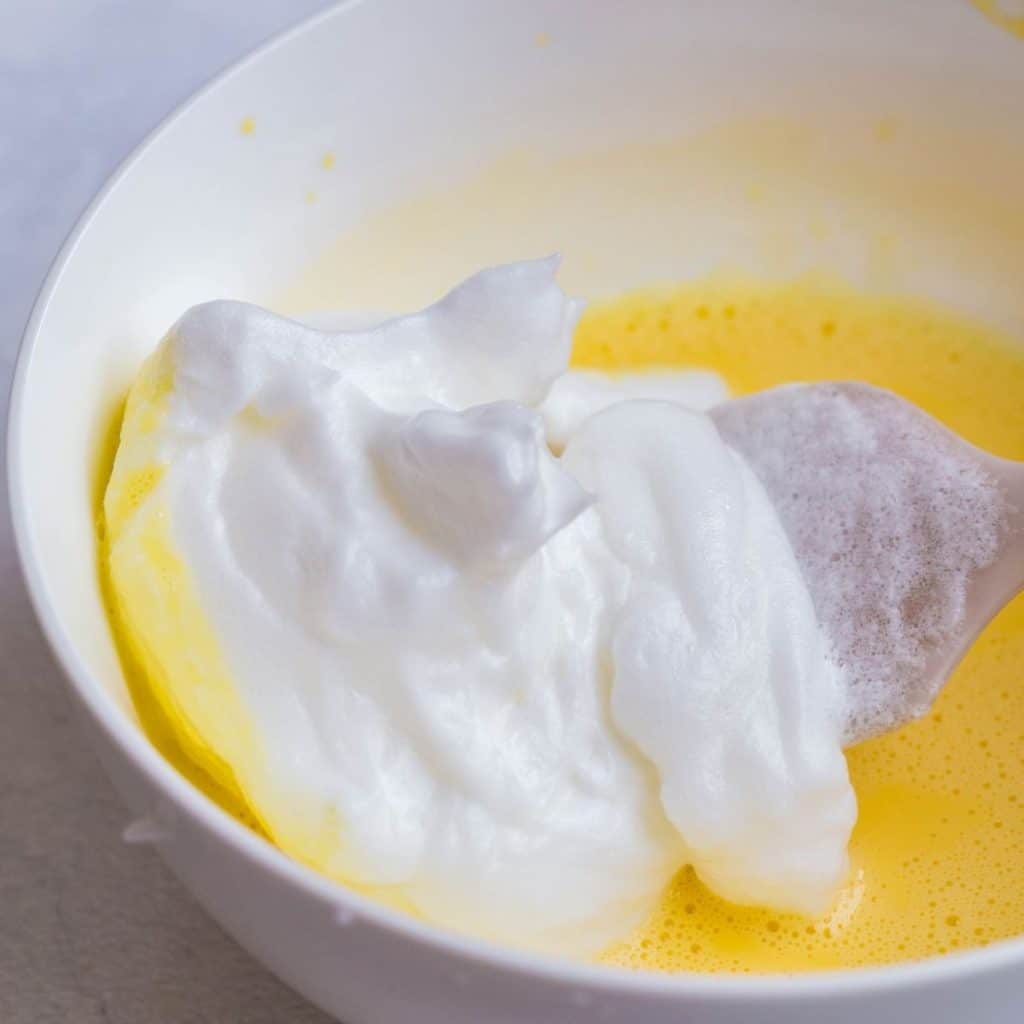 A white bowl with egg yolk mixture and egg white stiff peaks being folded together.