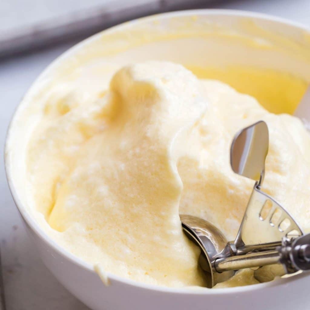 A white bowl with egg yolk mixture and egg white stiff peak mixture combined together and being scooped out with an ice cream scoop.