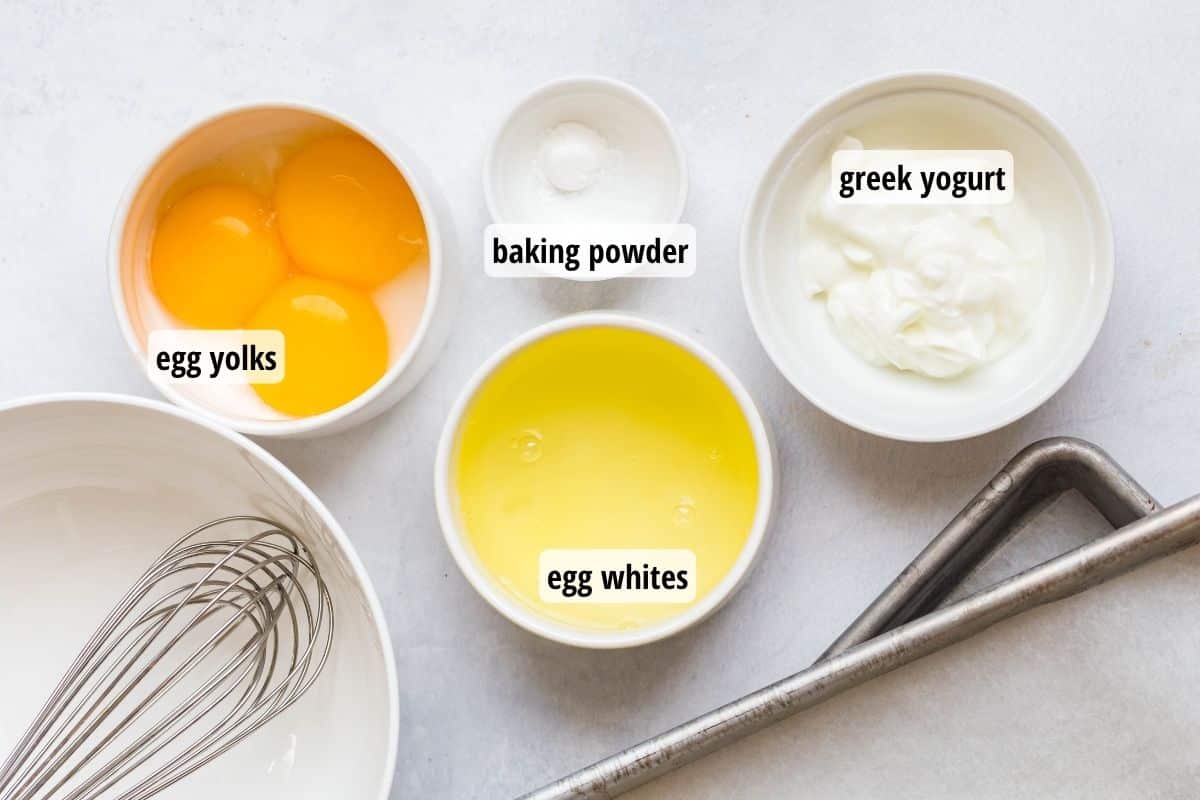 A white counter top with bowls full of egg yolks, egg whites, baking powder, greek yogurt and an empty bowl with a whisk inside of it.
