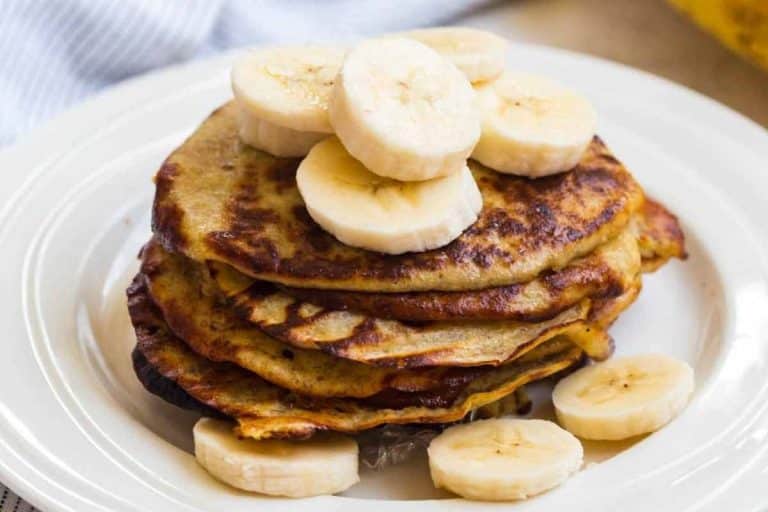Stack of banana pancakes on a white plate with sliced bananas on top and around the bottom of the stack.
