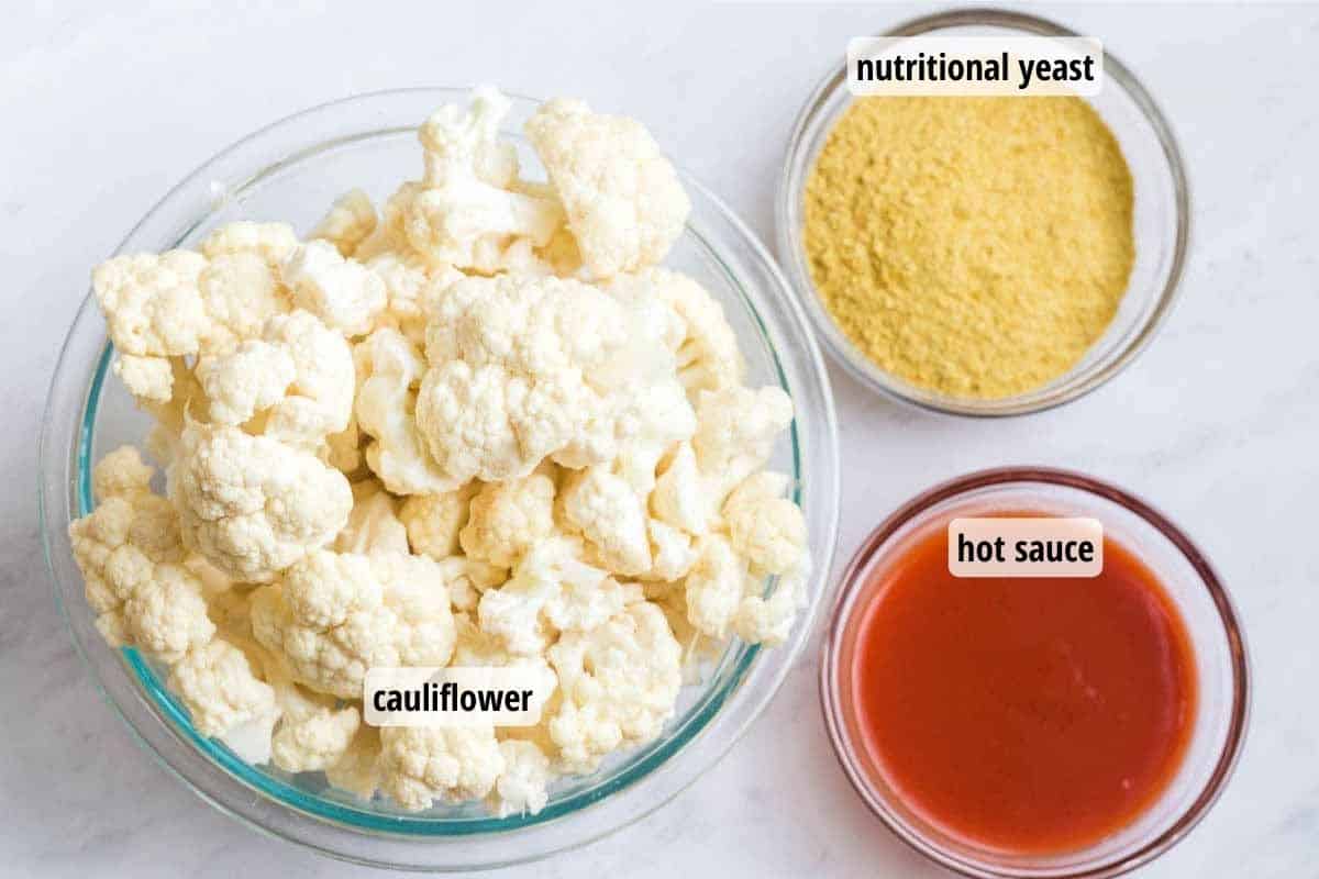 White counter with glass bowls containing cauliflower florets, nutritional yeast, and hot sauce.
