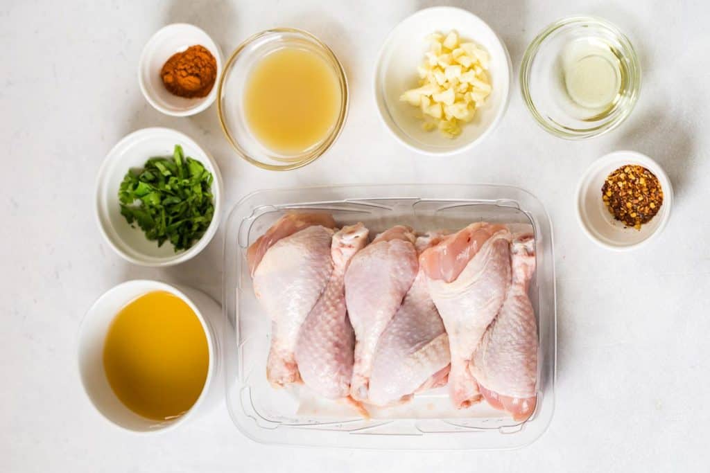 white counter with raw chicken legs and small bowls full of spices to make chicken recipe.