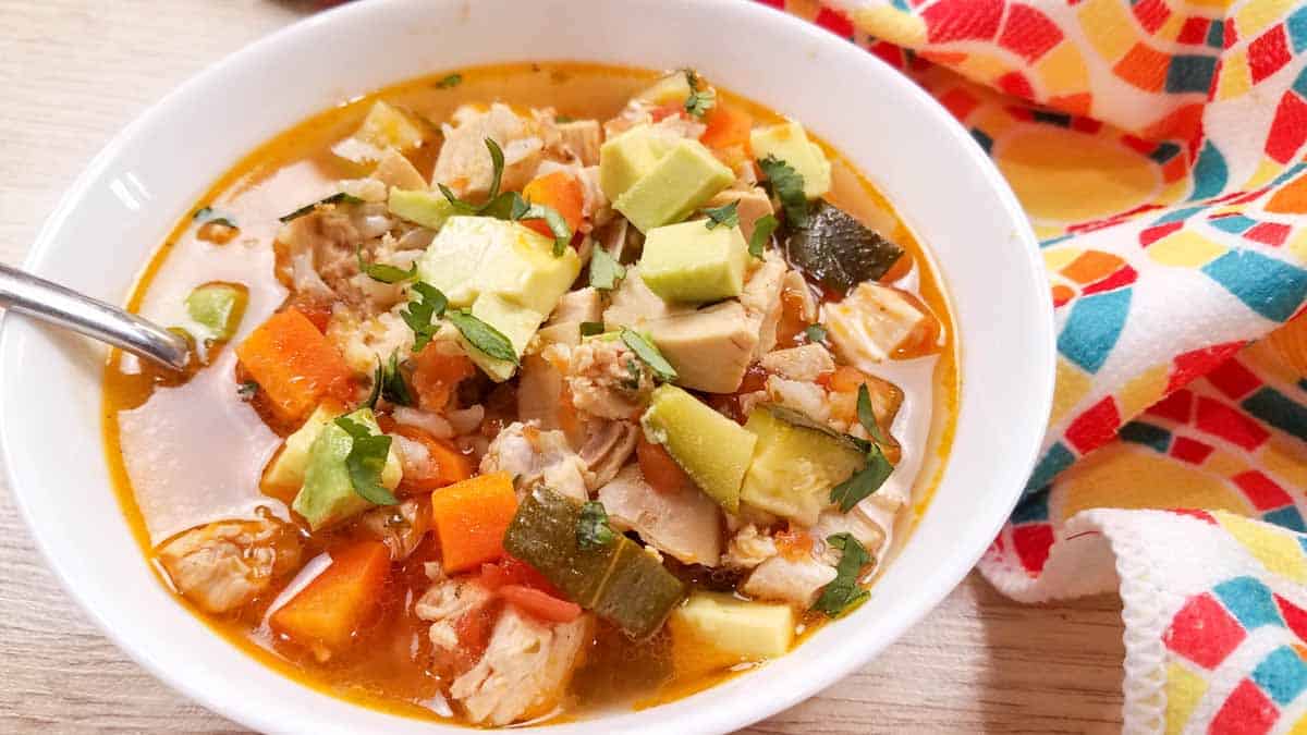 Rotisserie Chicken Soup in a white bowl with a spoon on top of a table with a colorful napkin.