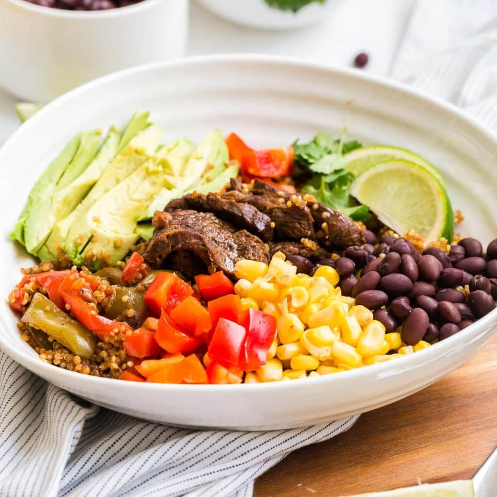 A white bowl of steak fajitas showing ingredients like sliced steak, bell peppers, corn, black beans, sliced avocado, quinoa, lime wedges, and cilantro.