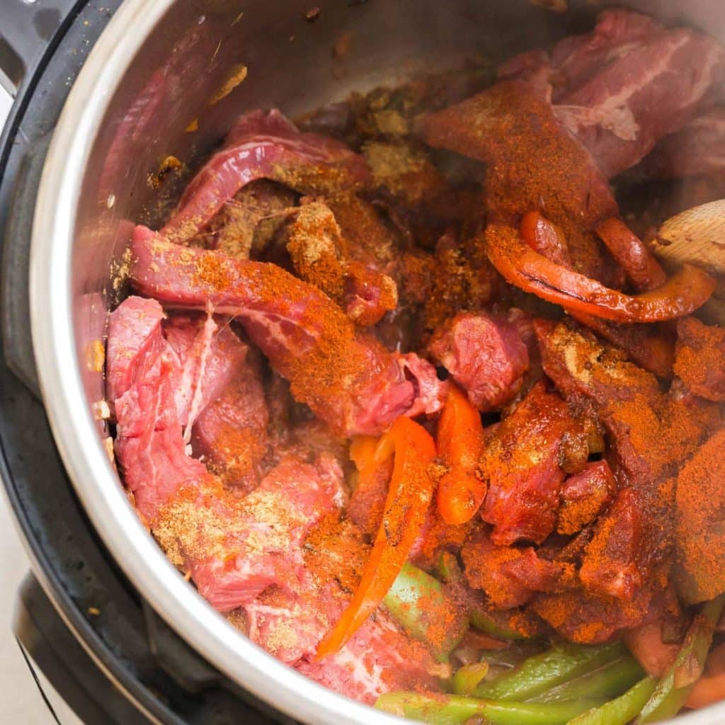 Inside an Instant Pot showing sliced steak with seasonings on top of fajita vegetables before being cooked.