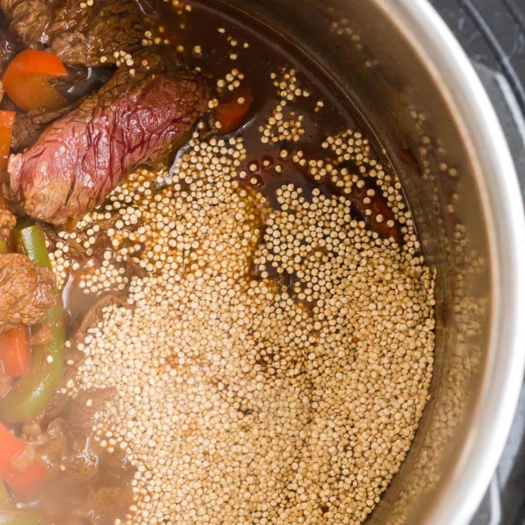 Inside an Instant Pot with fajita veggies and sliced steak covered with beef broth and topped with quinoa before being cooked.