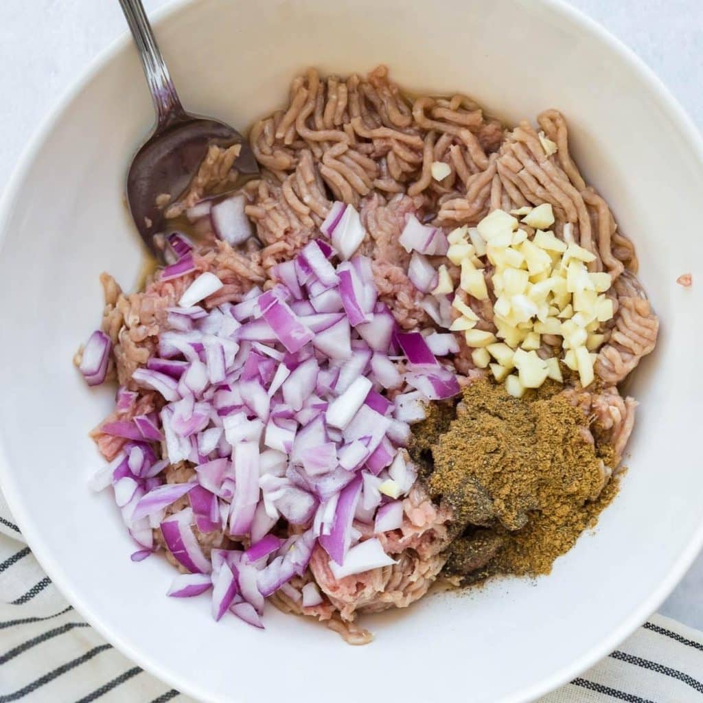 A white bowl with raw ground turkey, chopped red onion, diced garlic and seasonings - all about to be stirred to make sausage patties.