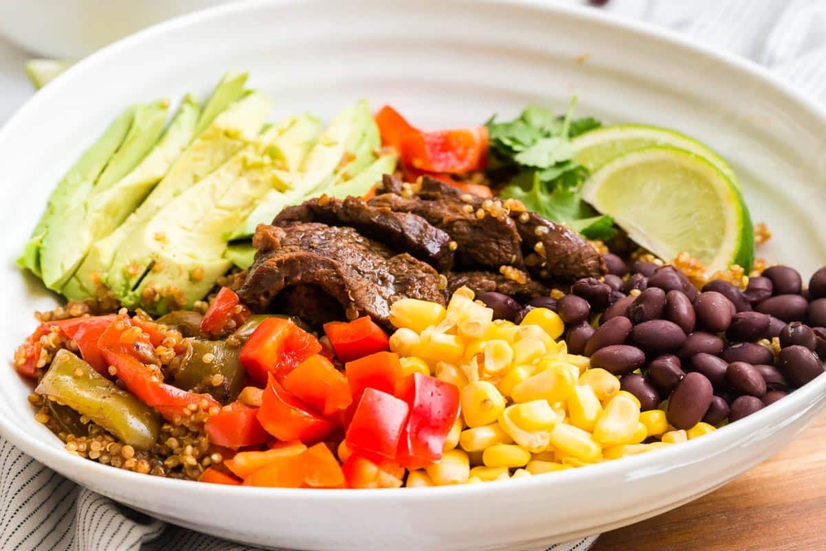 A white bowl of steak fajitas showing ingredients like sliced steak, bell peppers, corn, black beans, sliced avocado, quinoa, lime wedges, and cilantro.