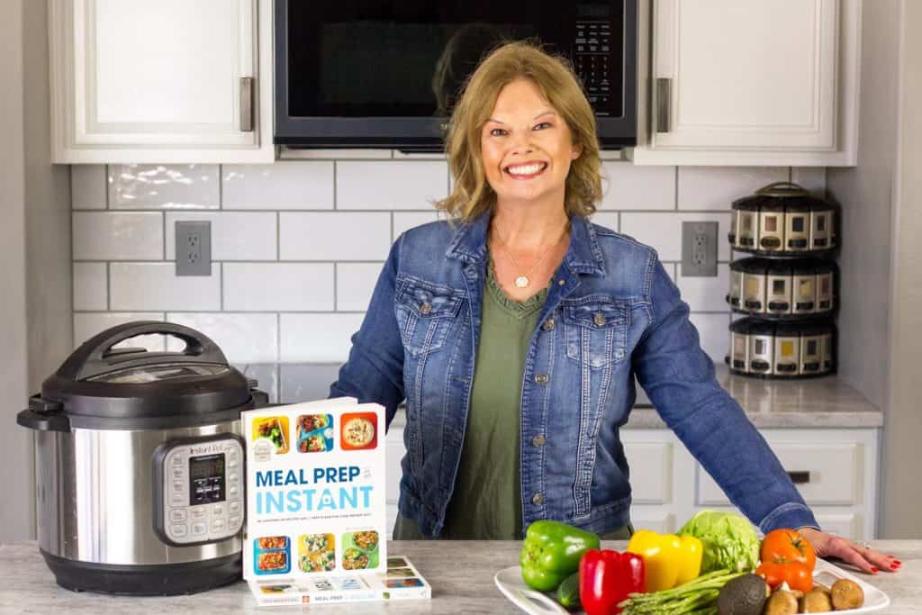Becca's wearing an olive green dress with a denim jacket and leaning on a white counter. Platter of veggies on the right side of the pic, with an Instant Pot and her book Meal Prep in an Instant on the left side of the pic. Microwave behind her along with white cabinets and subway tile backsplash.